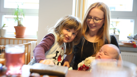 Eine ehrenamtliche Familienhelferin hat ein Baby auf dem Arm und das Geschwisterkind neigt sich zu ihr.