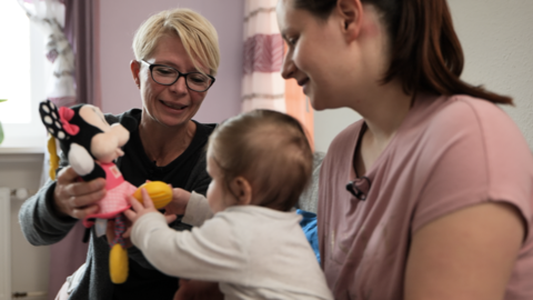 Eine Familienhebamme hat ein Kuscheltier in der Hand und sitzt mit einer Mutter und ihrem Baby auf dem Sofa der Familie.