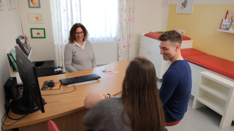 Ein junges Paar mit Baby sitzt einer Kinderärztin an einem Tisch gegenüber.