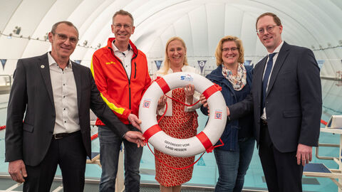 SWIMplus v.l.n.r.: Matthias Wörner (Vorsitzender Erster Offenbacher Schwimmclub von 1896 e.V.), Michael Hohmann (Präsident Deutsche Lebens-Rettungs-Gesellschaft Landesverband Hessen e.V. (DLRG), Sportministerin Diana Stolz, Juliane Kuhlmann (Präsidentin Landessportbundes Hessen (lsb h)) und Michael Scragg (Präsident Hessischer Schwimm-Verband e.V.) stehen in einer Schwimmhalle nebeneinander. Sportministerin Stolz hält einen Rettungsring vor sich und alle lächeln.