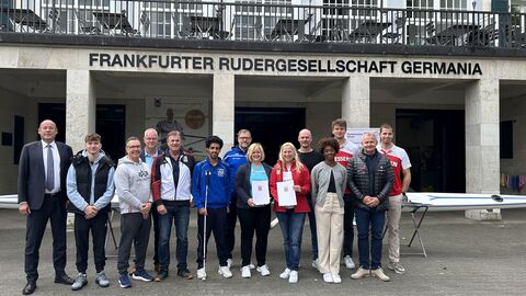 Sportministerin Diana Stolz hält Förderbescheide und steht mit mehreren Olympioniken vor dem Gebäude der Frankfurter Rudergesellschaft Germania