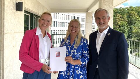 Sportministerin Stolz steht zwischen einer Frau und einem Mann. Sie übergibt der Dame links von sich einen Förderbescheid und alle lächeln in die Kamera.