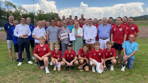 Sportministerin Diana Stolz steht mit einer Jugend Fußballmannschaft und Mitgliedern des Sportvereins auf einer Wiese und hält den Bescheid in der Hand.
