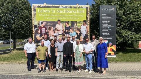 Gesundheitsministerin Diana Stolz steht inmitten einer Gruppe Personen auf einem Gehweg vor einem großen Aufsteller mit dem Titel "Leben in Nachbarschaft".