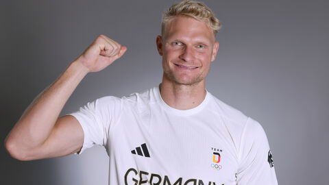 Ein blonder Mann mit weißem T-Shirt streckt den Arm hoch und hat einen begeisterten Gesichstausdruck