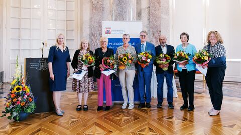 V.l.n.r.: Staatsministerin Diana Stolz, Anke Frieda Kraft, Anita Spieker, Ingrid Richber, Leonhard Helm, Uwe Heil, Greta Schrank und Susanne Zellmer Gruppenbild mit allen sieben Bürgerinnen und Bürgern. Sie stehen nebeneinander und halten einen Strauß Blumen in der Hand sowie die Pflegemedaille.