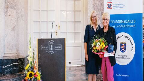Staatsministerin Diana Stolz und Anita Maria Spieker Zwei Frauen stehen neben einem Rednerpult. Eine hält einen Strauß Blumen in der Hand sowie die Pflegemedaille.