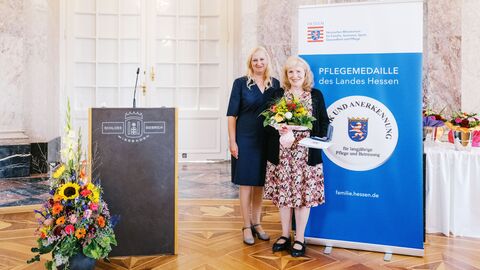 Zwei Frauen stehen neben einem Rednerpult. Eine hält einen Strauß Blumen in der Hand sowie die Pflegemedaille. Staatsministerin Diana Stolz und Anke Frieda Kraft.