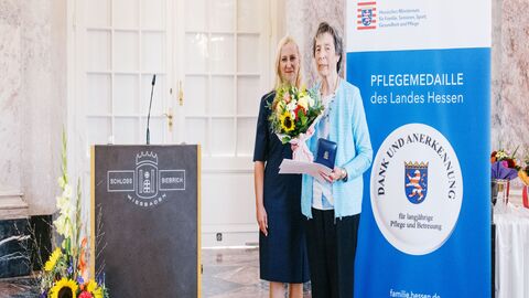 Staatsministerin Diana Stolz und Greta Schrank. Zwei Frauen stehen neben einem Rednerpult. Eine hält einen Strauß Blumen in der Hand sowie die Pflegemedaille.
