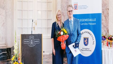 Staatsministerin Diana Stolz und Leonhard Helm. Eine Frau und ein Mann stehen neben einem Rednerpult. Der Mann hält einen Strauß Blumen in der Hand sowie die Pflegemedaille.