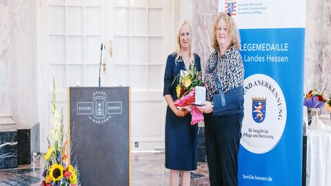 Staatsministerin Diana Stolz und Susanne Zellmer. Zwei Frauen stehen neben einem Rednerpult. Eine hält einen Strauß Blumen in der Hand sowie die Pflegemedaille.