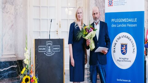 Staatsministerin Diana Stolz und Uwe Heil. Eine Frau und ein Mann stehen neben einem Rednerpult. Der Mann hält einen Strauß Blumen in der Hand sowie die Pflegemedaille.