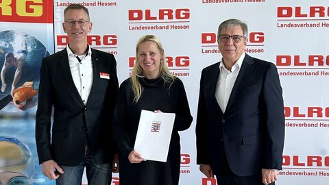 Präsident der DLRG Michael Hohmann, Sportministerin Diana Stolz und Oberbürgermeister Gert-Uwe Mende vor einem Poster der DLRG. Die Frau steht in der Mitte und hält den Bescheid in den Händen.