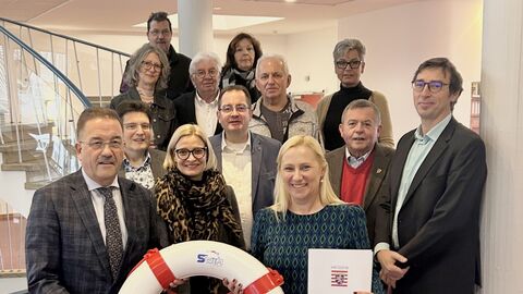 Mehrere Personen stehen hinter- und nebeneinander. Eine Frau und ein Mann halten einen Rettungsreifen in der Hand.
