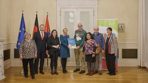 Mehrere Personen stehen vor 3 Flaggen und lächeln in Kamera. Eine Frau überreicht einem Mann eine Auszeichnung.