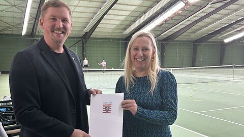 Ein Mann und eine Frau stehen in einer Tennishalle und halten lächelnd einen Förderbescheid in die Kamera