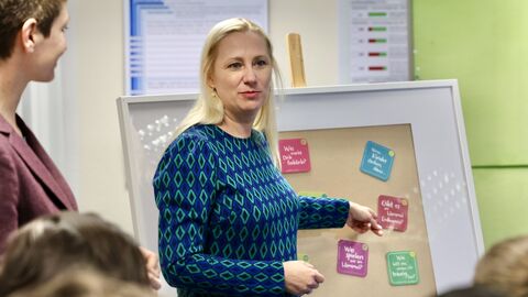 Familienministerin Diana Stolz steht vor einer Gruppe Personen und präsentiert einen Bilderrahmen, in dem bunte beschriebene Quadrate zu sehen sind.