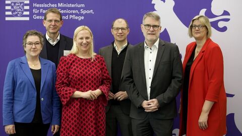 Eine kleine Gruppe Personen steht zum Gruppenfoto aufgestellt vor einem lila Aufsteller mit der Aufschrift "Hessisches Ministerium für Familie, Senioren, Sport, Gesundheit und Pflege".