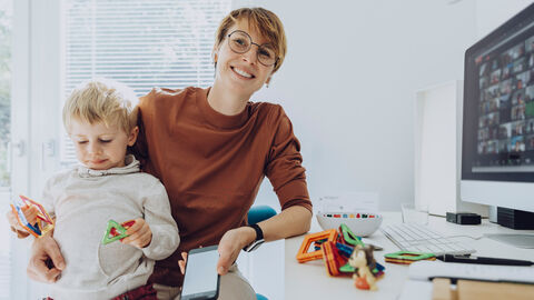 Eine Frau sitzt mit Kleinkind auf dem Schoß an ihrem Schreibtisch.