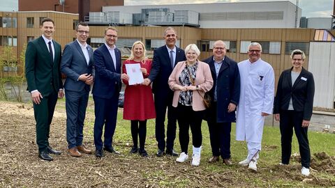 Eine Gruppe Personen steht auf einem Grundstück, im Hintergrund ist ein Gebäude zu erkennen. 