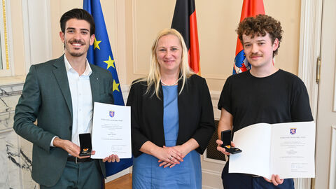 Gesundheitsministerin Stolz steht mit zwei Personen zusammen. Die beiden halten Urkunden in Händen.