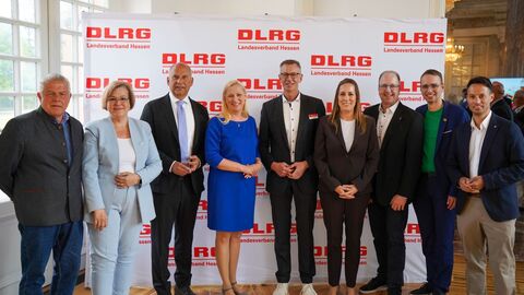 9 Menschen stehen nebeneinander und hinter ihnen eine Wand bedruckt mit der Aufschrift DLRG.