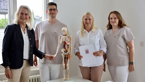 Vier Personen stehen in einem Raum, eine Person hält ein Dokument in den Händen, eine andere berührt ein kleines Skelett.