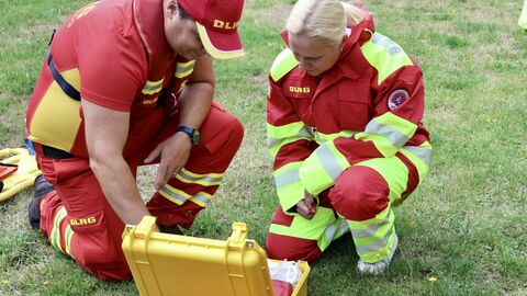 Ministerin packt beim Zusammenstellen des Einsatzmaterials an. Zwei Personen in DLRG Kleidung hocken auf der Wiese vor einer gelben Kiste.