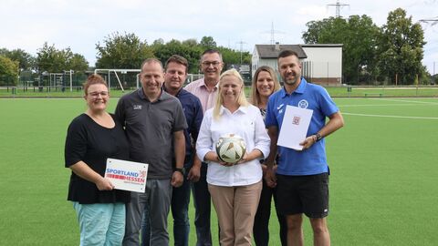 Eine kleine Personengruppe steht zur Überreichung des Förderbescheid auf einem Sportplatz.