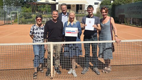 Die Sportministerin steht mit fünf weiteren Personen auf einem Tennisplatz und hält ein Schild mit "Sportland Hessen" in der Hand.
