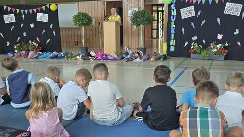 Kinder sitzen auf Matte in Turnhalle mit Schulranzen.