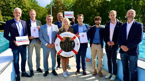 Neun Personen stehen in einem Freibad zwischen zwei Schwimmbecken nebeneinander. Die mittlere Person hält einen Rettungsring hoch.