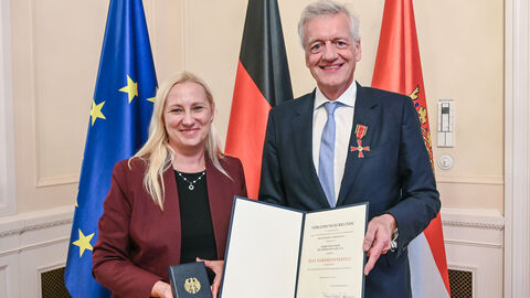 Diana Stolz hält einen Verdienstorden in den Händen und steht neben Prof. Dr. Ferdinand M. Gerlach, der eine Urkunde präsentiert. Hinter ihnen sind die Flaggen von Europa, Deutschland und Hessen zu sehen.