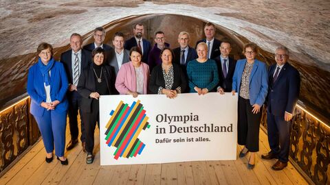Eine Gruppe von 15 Personen steht in einem Kellergewölbe - vor Ihnen ein Schild mit der Aufschrift "Olympia in Deutschland".