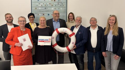 Neun Personen stehen nebeneinander in einem Raum. Eine hält eine Urkunde hoch und daneben jemand einen weißen Schwimmring mit roten Streifen.