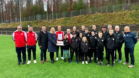 Eine Gruppe von Erwachsenen und Kindern, teils in Vereinskleidung, stehen auf einem Rasen-Fußballfeld und schauen in die Kamera.
