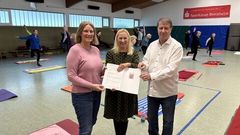 Drei Menschen stehen in einer Sporthalle. Im Hintergrund sind Sportmatten ausgelegt.
