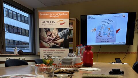 Ein Laptop sowie Gläser und Brillen liegen auf einem Tisch. Im Hintergrund ist ein Aufsteller mit der Aufschrift "Auxilium Hospizverein Wiesbaden".