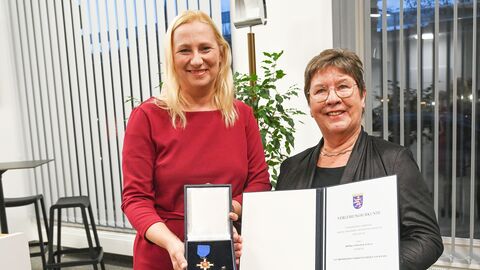 Gesundheitsministerin Stolz und Frau Schulz stehen nebeneinander in einem Raum und halten die Urkunde sowie den Orden hoch.