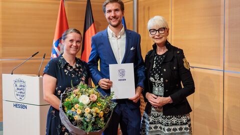 Drei Personen stehen vor den Flaggen von Hessen, Deutschland und der EU. Die Frau links hält einen Blumenstrauß, der Mann in der Mitte eine Urkunde.