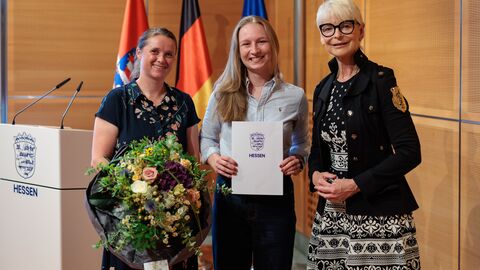 Drei Personen stehen vor den Flaggen von Hessen, Deutschland und der EU. Die Frau links hält einen Blumenstrauß, die Frau in der Mitte eine Urkunde.