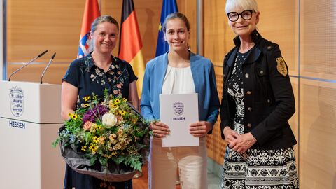 Drei Personen stehen vor den Flaggen von Hessen, Deutschland und der EU. Die Frau links hält einen Blumenstrauß, die Frau in der Mitte eine Urkunde.