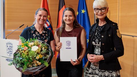 Drei Personen stehen vor den Flaggen von Hessen, Deutschland und der EU. Die Frau links hält einen Blumenstrauß, die Frau in der Mitte eine Urkunde.