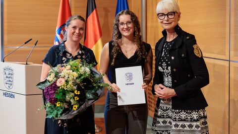 Drei Personen stehen vor den Flaggen von Hessen, Deutschland und der EU. Die Frau links hält einen Blumenstrauß, die Frau in der Mitte eine Urkunde.