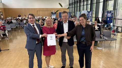 Vier Personen, darunter auch Gesundheitsministerin Stolz, stehen nebeneinander in einer Halle und halten eine Urkunde hoch.