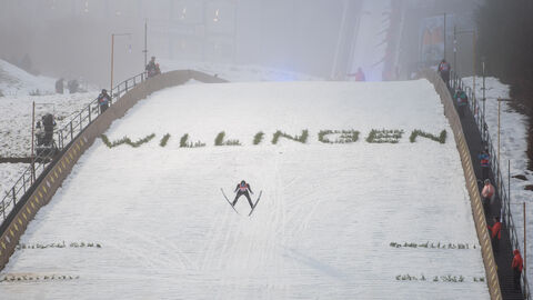 Skispringen WIllingen Skispringer springt über Skischanze auf der das Wort "Willingen" in Gras geschrieben ist.