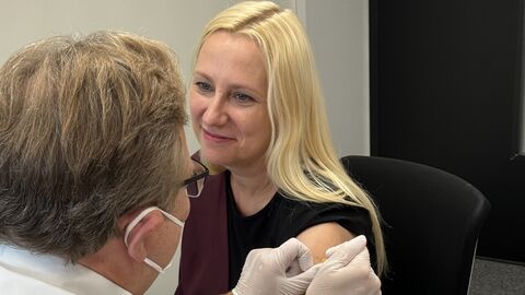 Gesundheitsministerin Stolz sitzt auf einem Stuhl. Ein Arzt ist gerade dabei ein Pflaster auf ihren Oberarm zu kleben.